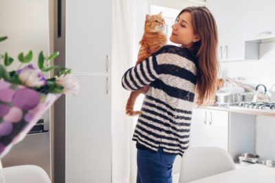 Young woman playing with cat in kitchen at home. Girl holding and raising red cat